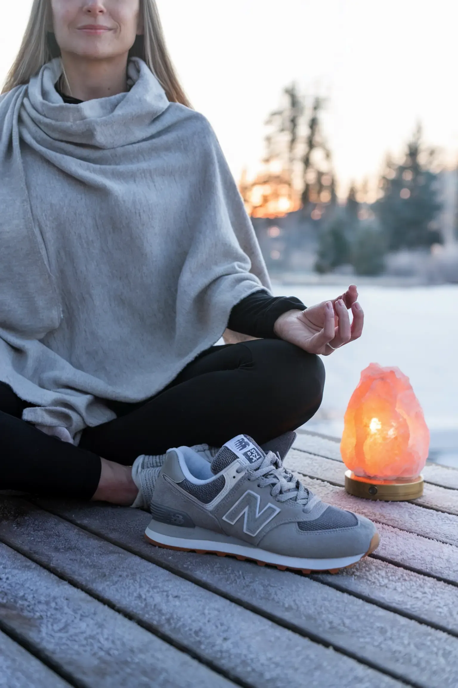 Woman in grey cowl neck sweater and black leggings practicing meditation on a winter deck, wearing grey New Balance sneakers with a Himalayan salt lamp nearby