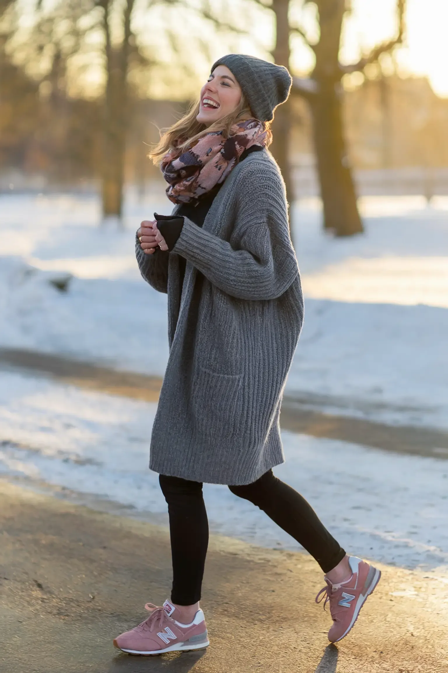 Woman laughing in grey oversized cardigan and patterned scarf during sunrise in snowy park, wearing dusty rose New Balance sneakers while holding coffee cup