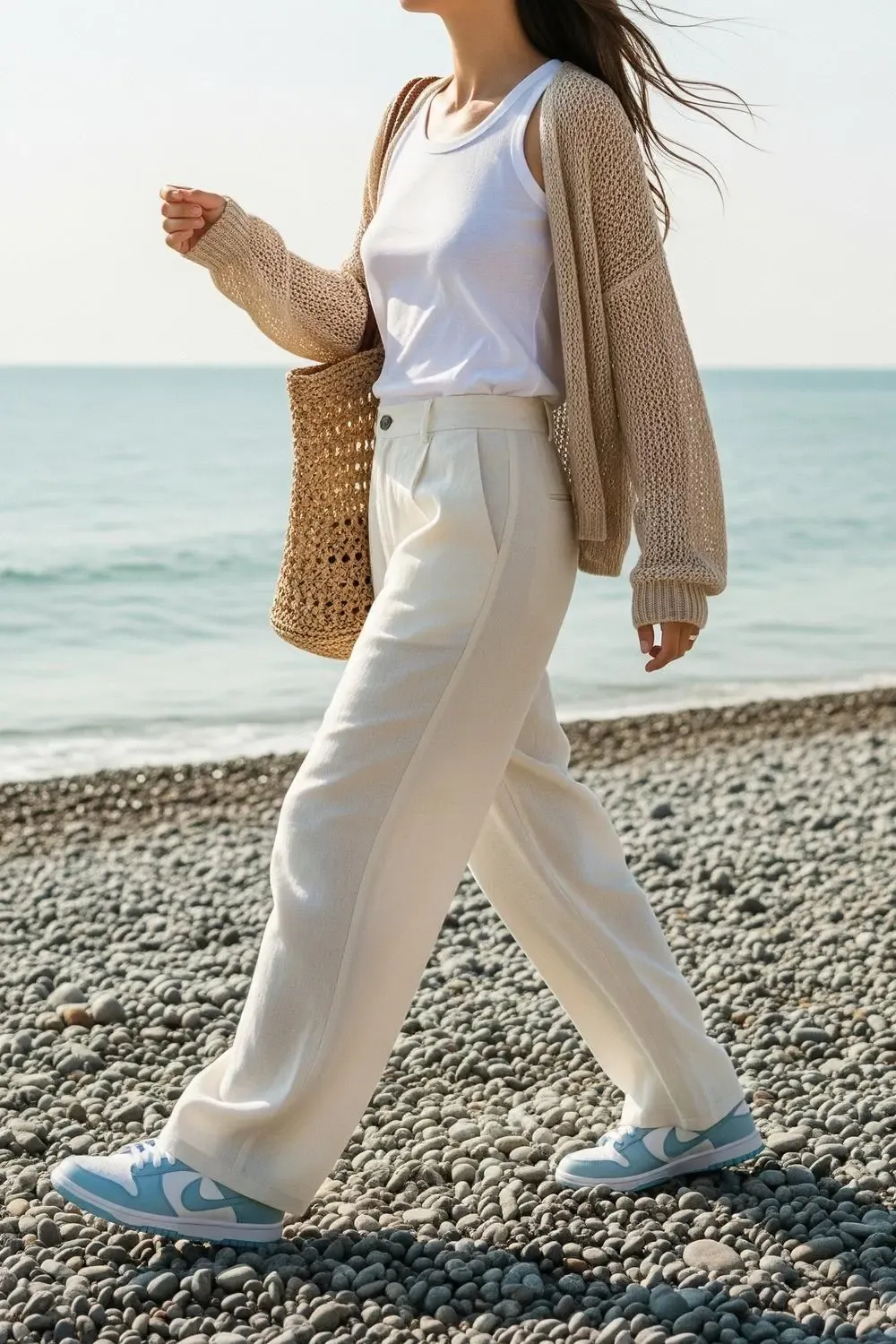 A woman exuding coastal elegance in wide-leg cream linen pants, a white tank top, a sandy-colored open-knit cardigan, and Nike Dunk Low 'UNC' sneakers, walking along a pebble beach.